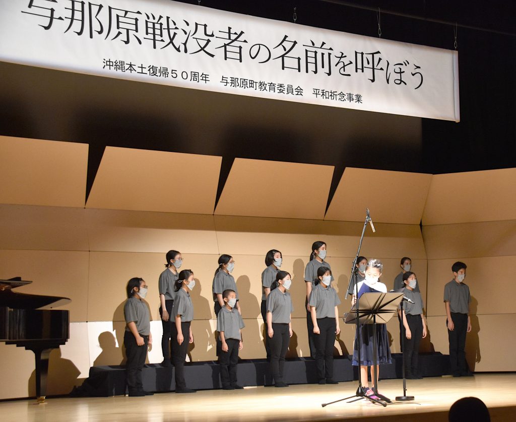 舞台上中央で戦没者名を読み上げる小学生の女の子。その背後には「うんたま合唱団11人が静かに合唱している
